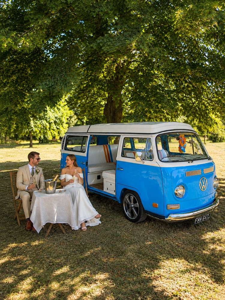 Bride and groom seated inside Mabel, toasting with champagne flutes, their bouquet on the seat beside them and greenery visible through the open door.