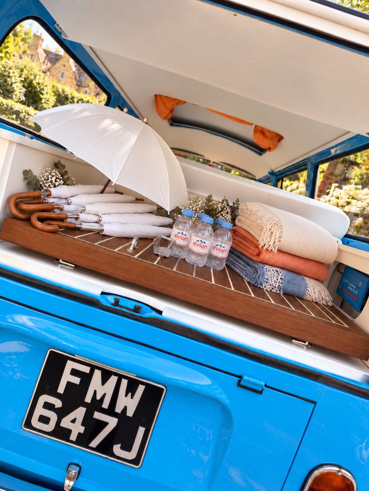 The open rear of Mabel with the picnic board down: a row of white wooden-handled umbrellas, chilled bottled water with glasses, and folded blankets in cream, blue and orange.