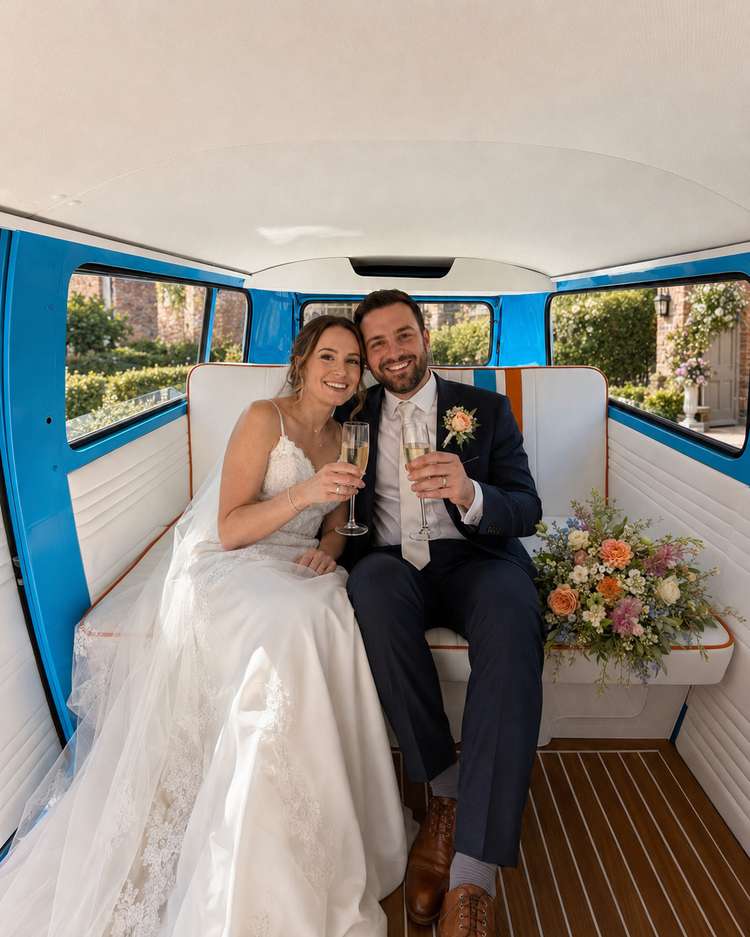 Bride and groom sharing a champagne toast at a table beside Mabel, parked in dappled shade with her side door open onto the striped vintage interior.